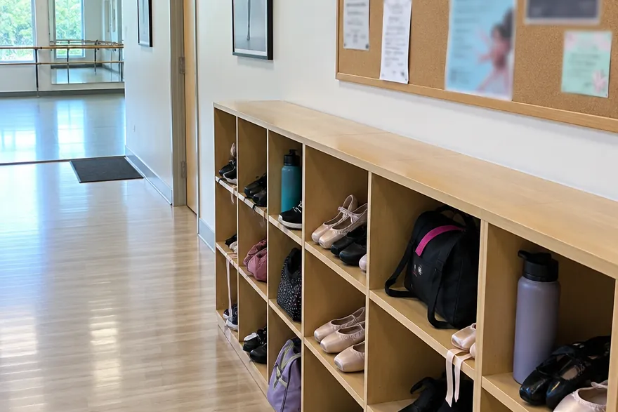 A dance studio hallway with wooden cubbies full of ballet shoes, bags and water bottles, a notice board above, and a studio with a barre at the end.