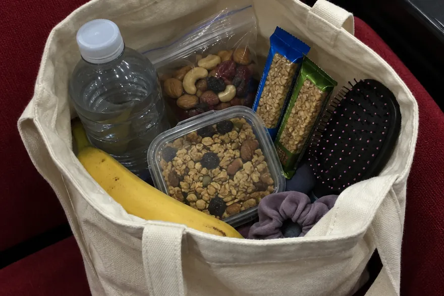 A canvas tote on a red auditorium seat holding a water bottle, nuts, granola bars, a banana and a hairbrush.